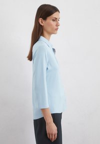 Light blue button-up shirt, made of soft fabric, features a collar and three-quarter sleeves, with a relaxed fit and no visible patterns.