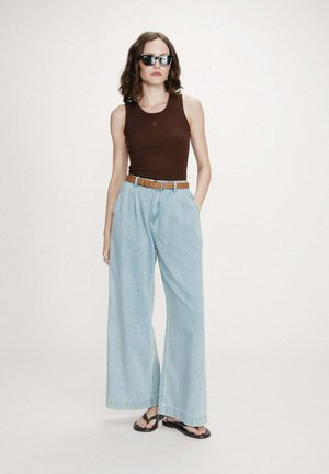 Femme portant des lunettes de soleil noires, un haut marron sans manches, un jean bleu clair à jambes larges avec une ceinture marron, et des sandales noires, debout devant un fond uni.