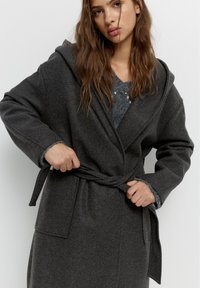 Grijze, lange wikkeljas met een capuchon, voorzien van een strikceintuur en zijzakken. Zacht materiaal met een gladde textuur en contrasterende binnenmanchetten.