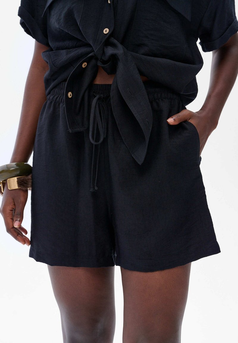 Zwarte linnen shorts met een elastische tailleband en koord. Voorzien van zijzakken en een losse pasvorm, gecombineerd met een geknoopt zwart topje.