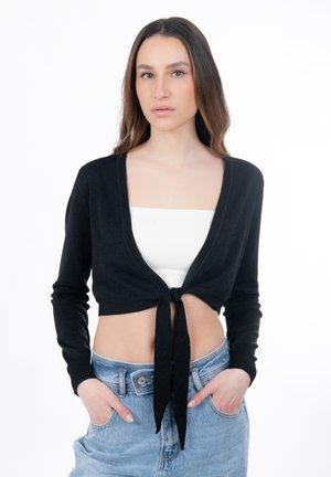 Jeune femme aux cheveux longs portant un gilet noir court noué sur le devant par-dessus un top bandeau blanc et un jean bleu clair, les mains dans les poches, expression neutre.