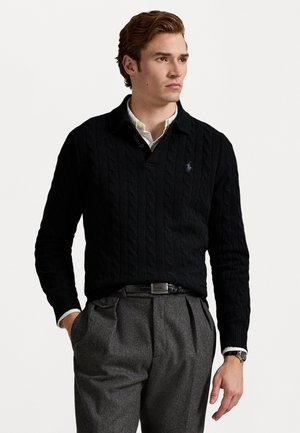 Zwarte kabelgebreide trui met een kraag, voorzien van een klein logo op de borst. Gestyled met grijze geplooide broek en een wit overhemd eronder.