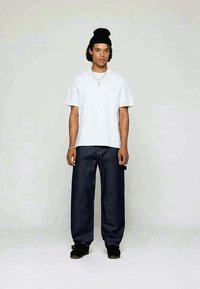Weißes Baumwoll-T-Shirt, lockerer Schnitt, kombiniert mit dunkelblauen, lässigen Jeans mit weitem Bein. Ein schwarzer Beanie und schwarze Schuhe runden das Outfit ab.