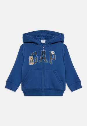 Sweat à capuche bleu pour tout-petit avec fermeture éclair à l'avant, grandes lettres "GAP", petit motif ours en peluche et poches kangourou à l'avant.