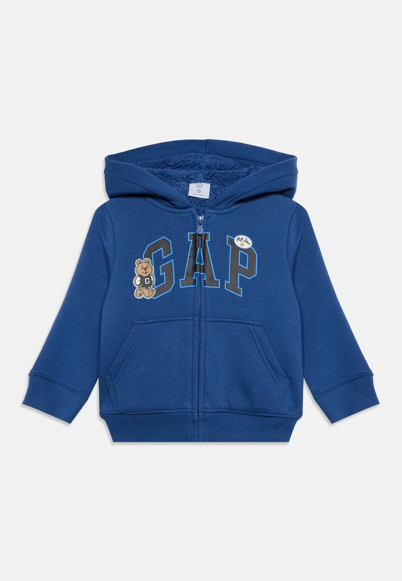 Sweat à capuche bleu pour tout-petit avec fermeture éclair à l'avant, grandes lettres "GAP", petit motif ours en peluche et poches kangourou à l'avant.