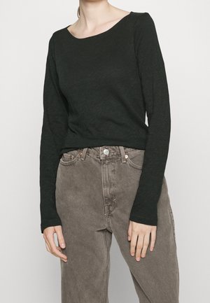 Vrouw die een strak zwart shirt met lange mouwen draagt, ingestopt in hooggetailleerde, losvallende grijze spijkerbroek, staand tegen een effen achtergrond.