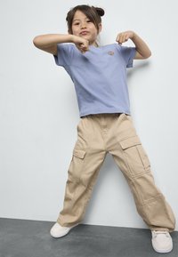 Camiseta de manga corta azul claro con un pequeño logo, combinada con pantalones cargo beige que tienen múltiples bolsillos y un ajuste relajado. Zapatillas blancas.