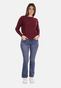 Maglione in maglia borgogna con texture a coste, scollo rotondo e maniche lunghe, abbinato a jeans azzurri e sneakers bianche.