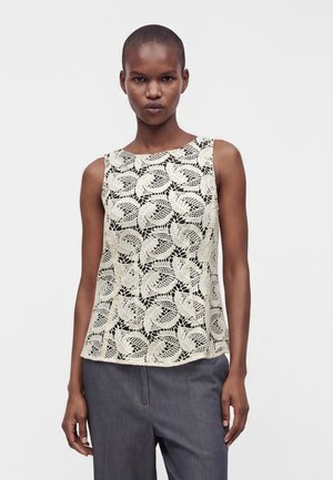 Femme aux cheveux courts portant un haut en dentelle blanche sans manches avec un motif de feuilles et un pantalon gris sur un fond clair uni.