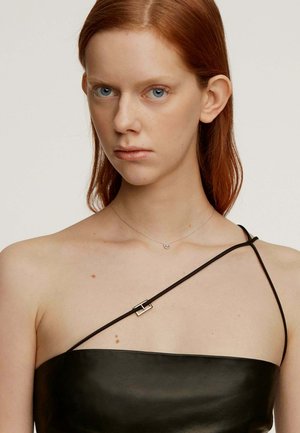 Top de cuero negro con una sola tira, que presenta un detalle de hebilla cuadrada plateada. La modelo tiene el cabello largo y rojo, y lleva un collar plateado sencillo.