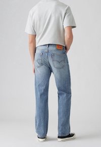 Person som bär ljusblå raka jeans, grå kortärmad tröja och svarta låga sneakers, står med händerna bakom ryggen.