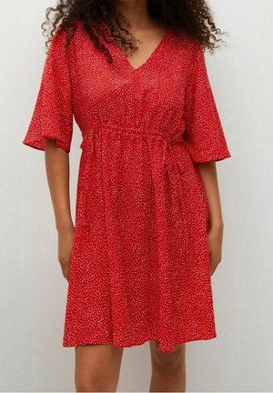Femme portant une robe rouge longueur genou avec de petits pois blancs et des manches mi-longues, debout sur un fond clair uni.