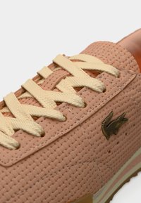 Baskets rose clair avec empeigne texturée, lacets jaunes et un logo crocodile doré sur le côté. Elles disposent d'une semelle en caoutchouc avec un design léger.