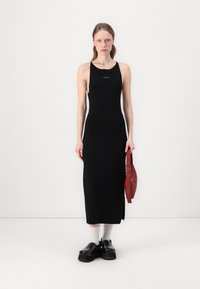 Abito midi senza maniche nero in tessuto liscio, con uno spacco laterale. La modella tiene in mano una borsa rossa, indossando scarpe platform nere chunky e calze bianche.