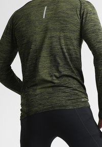 Långärmad träningströja i mörkgrön med ett strukturerat mönster, reflekterande detaljer på ryggen och en åtsittande passform.
