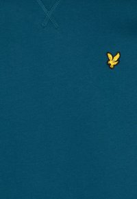 T-shirt in cotone color teal con scollatura a V e colletto a costine, dotato di un piccolo logo di un'aquila dorata ricamato sul lato sinistro del petto.