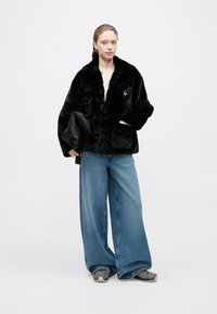 Chaqueta de pelaje sintético negro con cuello y dos bolsillos frontales, combinada con unos vaqueros de mezclilla azules de corte suelto y zapatillas grises, llevada con un bolso negro.