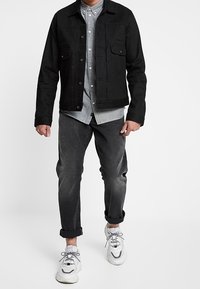 Chaqueta de mezclilla negra con dos bolsillos en el pecho, camisa gris abotonada, jeans grises desgastados y zapatillas blancas con detalles en gris.
