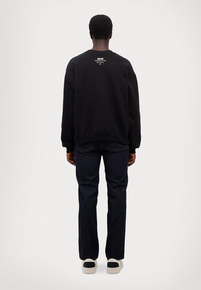 Felpa nera a girocollo con dettaglio del logo sul retro, abbinata a jeans di denim blu scuro e sneakers chiare, vista da dietro.