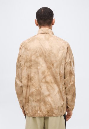 Άνδρας που φοράει ένα ανοιχτό καφέ, μπουφάν με σχέδιο ταϊ-ντάι και το κείμενο "TERREX" στο αριστερό μανίκι, στέκεται γυρισμένος με την πλάτη προς τα εμπρός, πάνω σε λευκό φόντο.