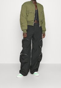 Olijfgroene bomberjack met twee ritszakken, gecombineerd met zwarte cargo-broeken met grote ritszakken en dik materiaal. Witte sneakers met groene accenten.