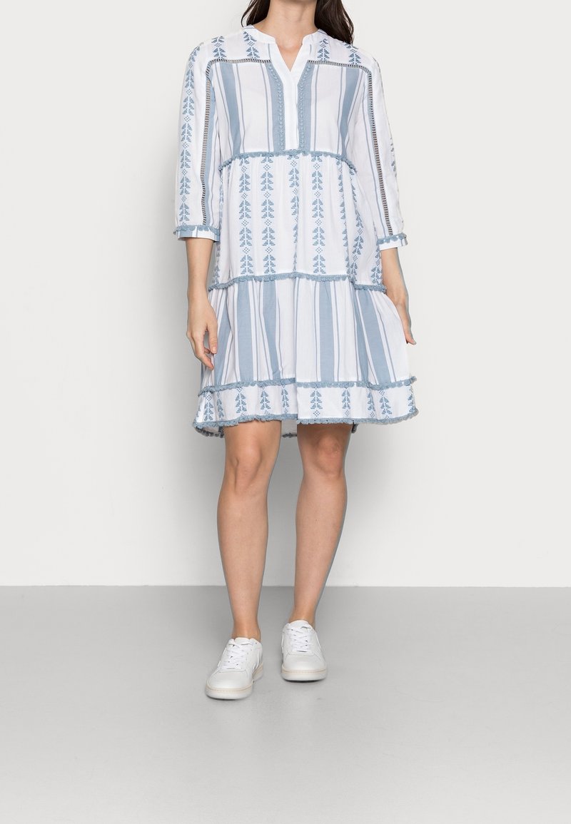 Biała i niebieska sukienka w paski z rękawami do łokcia, o warstwowym kroju i haftowanymi detalami. Połączona z białymi trampkami.