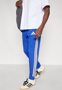 Kráľovsky modré tepláky s tromi zlatými vertikálnymi pruhmi, elastickými manžetami a logom Adidas na stehne, doplnené čiernymi a krémovými teniskami.