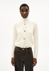 Cardigan crema con scollatura rotonda, cinque bottoni e polsini a costine, abbinato a jeans scuri con vita regolabile. Tessuto morbido.
