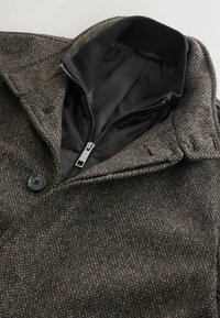 Brun texturerad ullkappa med svart innerfoder, dragkedjeförslutning och två knappar fram. Har krage och detaljerade sömnadsdetaljer.