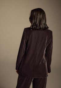 Ciemny aksamitny blezer z długimi rękawami, gładką fakturą i prostym krojem. Tył zdobi prosty szew bez ozdób.