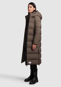 Lång, pösig brun dunjacka med huva, sidodragkedjor och svarta detaljer. Stiliserad med svarta knähöga stövlar och slim fit byxor.