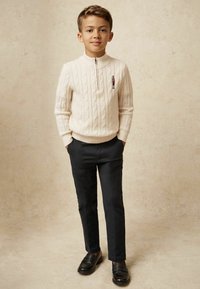 Pull en crème tricoté avec un col zippé mi-hauteur, associé à un pantalon noir ajusté et des mocassins noirs. Le pull est orné d'une petite figure brodée.