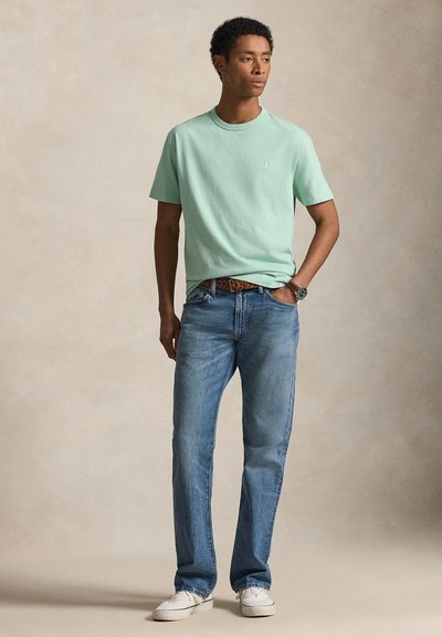 T-shirt de manga curta verde menta, jeans de ganga azul claro, cinto castanho trançado, ténis brancos, em pé com as mãos nos bolsos contra um fundo neutro.