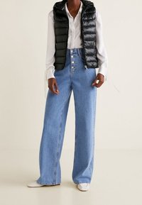 Černá péřová vesta s kapucí přes bílou košili, doplněná světle modrými širokými džínami a bílými nazouvacími botami.