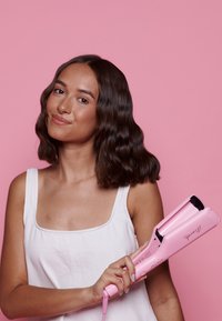 Herramienta de peinado de color rosa con bordes redondeados y un mango texturizado, sostenida por una persona. El fondo es de color rosa sólido y el producto presenta el logo de la marca.