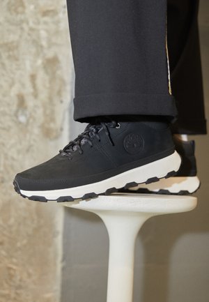 Schwarze Schnürsneaker mit Gummisohle, Lederoberteil und Logo an der Seite, präsentiert auf einem weißen Sockel vor einer strukturierten Wand.