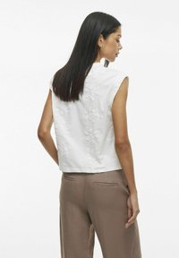 Top sin mangas blanco con patrón floral bordado, dobladillo redondeado y textura sutil. Combinado con pantalones de pierna ancha de color marrón claro.