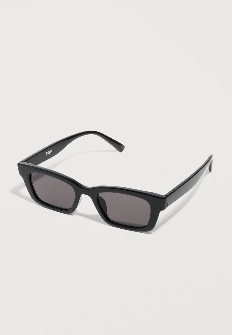 Μαύρα ορθογώνια γυαλιά ηλίου με χοντρά πλαίσια και σκουρόχρωνους φακούς που ακουμπούν σε λευκή επιφάνεια.