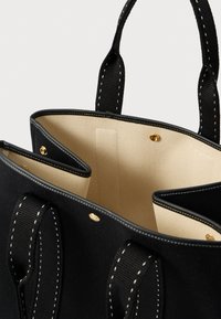 Μαύρη καμβάς τσάντα tote με χρυσές λεπτομέρειες, διαθέτοντας μπεζ επένδυση και λευκές ραφές. Το εσωτερικό διαθέτει πολλαπλές θήκες και ανθεκτικά χερούλια.