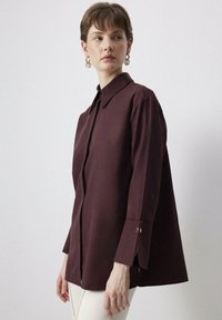 Camicia button-up bordeaux con taglio dritto, colletto, maniche lunghe e spacchi laterali. Tessuto liscio con una leggera lucentezza.