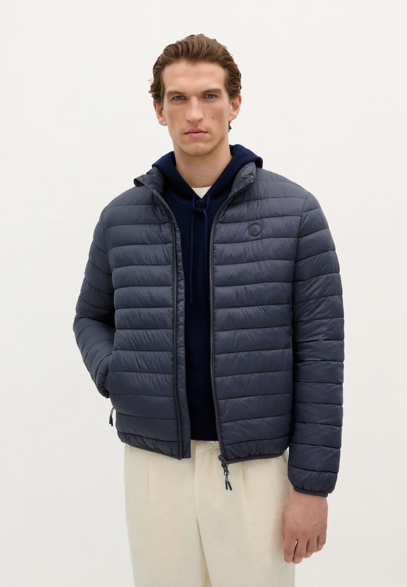 Gevoerde jas in donkergrijs, met een rits aan de voorkant, taps toelopende manchet en een capuchon; gedragen over een donkerblauwe hoodie en lichtbeige broeken.