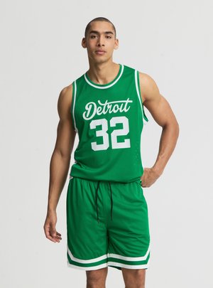 Uomo in piedi che indossa maglietta e pantaloncini da basket verdi con la scritta "Detroit" e il numero 32 in bianco sulla maglietta, mani rilassate sui fianchi.