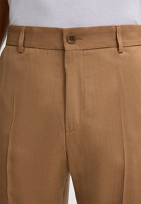 Close-up of brown pants with belt loops, a buttoned waistband, and a white tucked-in shirt visible at the top.