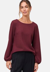Pull en velours de couleur bordeaux avec un col rond et des manches longues et volumineuses, associé à un pantalon noir.