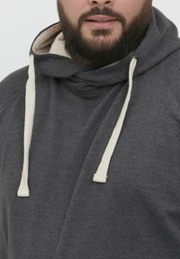 Sudadera gris con cuello tipo cowl, con cordones en color crema, textura suave y un corte holgado. El material parece similar al forro polar y es cálido.