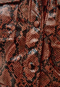 Veste en cuir synthétique brun avec un motif texturé de serpent, présentant des accents noirs et des boutons ronds.