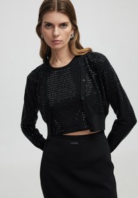 Schwarze kurze Strickjacke mit strukturiertem, offenem Gewebe, langen Ärmeln und Knopfverschluss. Kombiniert mit einem figurbetonten schwarzen Rock.