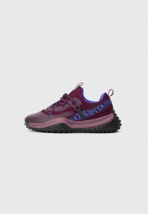 Burgundy athletic sneaker with black bumpy sole, blue interior, adjustable elastic laces, and blue "Versace Jeans Couture" text on side strap.
