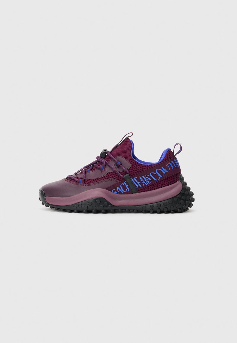 Burgundy athletic sneaker with black bumpy sole, blue interior, adjustable elastic laces, and blue "Versace Jeans Couture" text on side strap.
