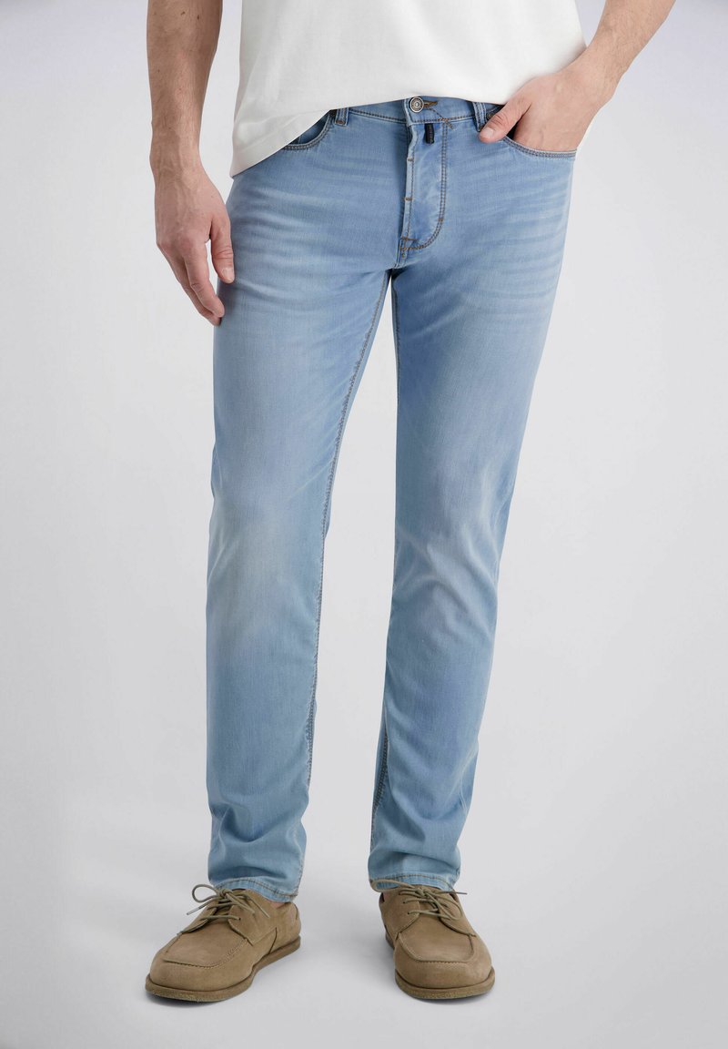 Mann trägt hellblaue, eng anliegende Jeans, beige Schnürschuhe und ein weißes Hemd, mit einer Hand in der Tasche und steht vor einem schlichten Hintergrund.